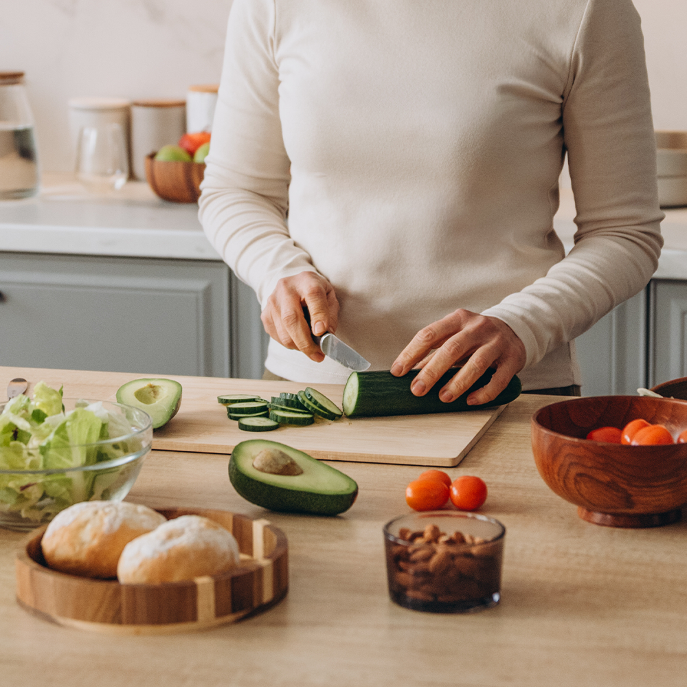 Femme qui découpe des légumes avec des aliments bon pour la santé tout disposés tout autour (amandes, avocats, tomates...)