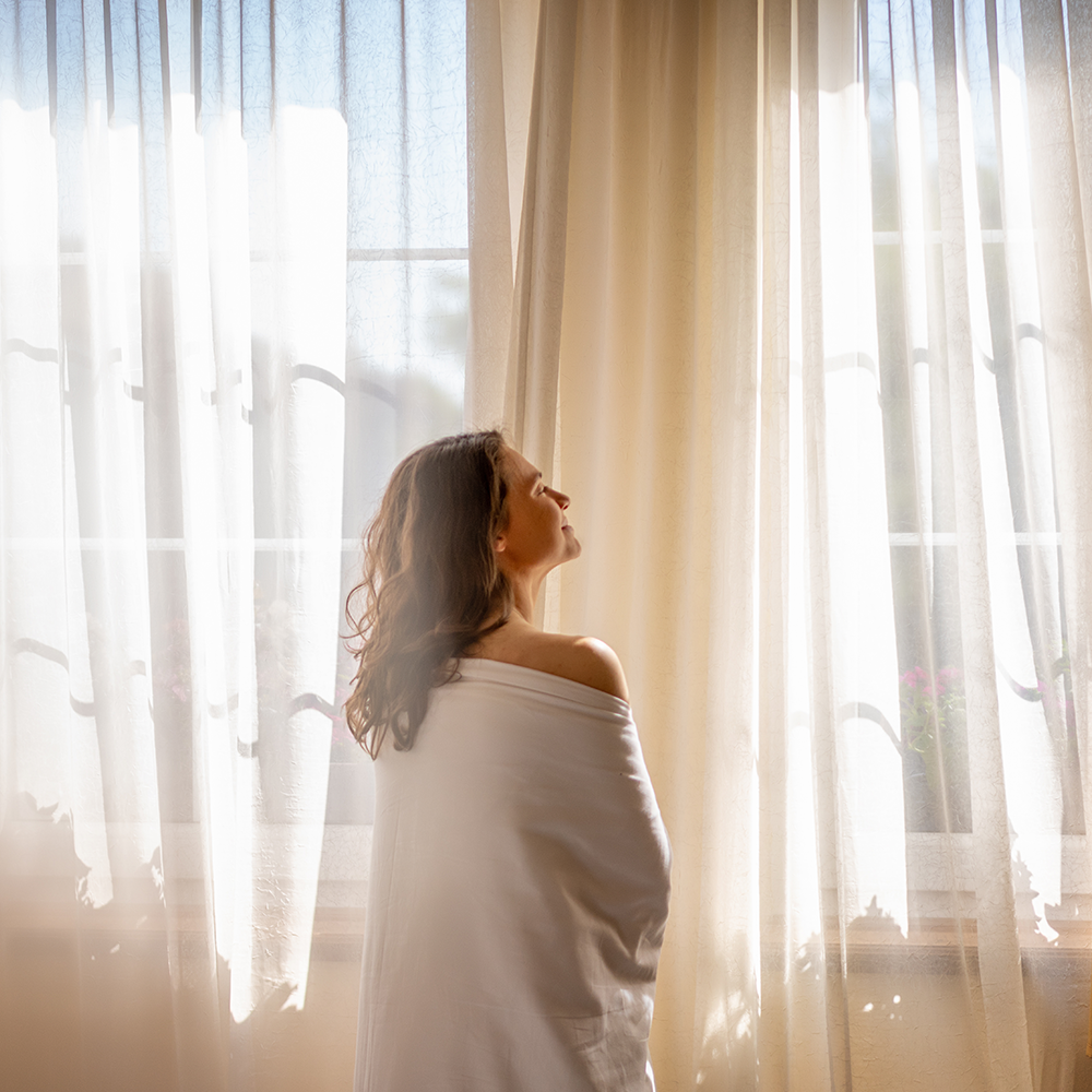 Jeune femme devant une fenêtre avec des rideaux transparents prenant le soleil